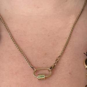 May include: Gold-coloured necklace with a decorative clasp set with green gemstones. The chain has a wheat-link design. The necklace is shown against a skin-toned background.