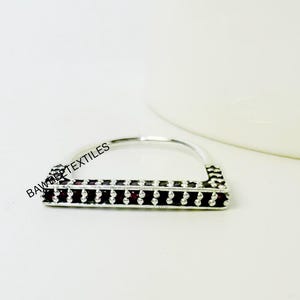 May include: A silver-colored ring with a rectangular design, featuring a row of small, dark-colored stones. The ring has a curved band and a geometric pattern on the edges. The ring is set against a white background.