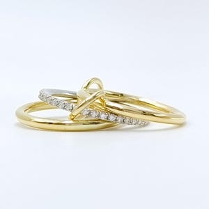 Op de afbeelding: Drie gouden ringen met een minimalistisch design. Eén ring is een simpele band, terwijl de andere twee in elkaar verweven zijn, één met een rij kleine, heldere stenen. De ringen staan tegen een witte achtergrond.