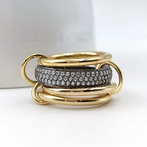 Op de afbeelding: Een goudkleurige ring met meerdere in elkaar grijpende banden. Eén band is bezet met kleine, sprankelende stenen. De ring staat tegen een witte achtergrond met een wit keramisch object op de achtergrond.