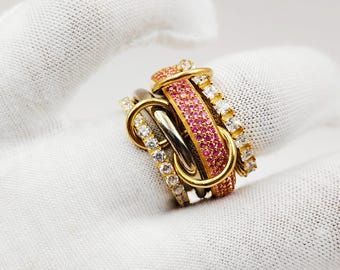 May include: A collection of gold, silver, and pink gemstone rings. The rings are stacked together, with some featuring clear and pink gemstones. The rings are displayed on a white surface.