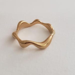 Puede incluir: Un anillo dorado con un diseño ondulado y orgánico. El anillo tiene un acabado liso y pulido y una forma irregular y fluida. La joya está sobre un fondo blanco.