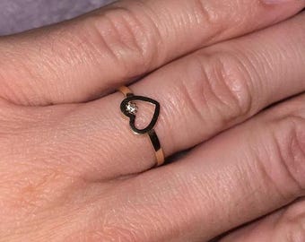 May include: A gold-colored ring featuring a heart-shaped design with a small, clear gemstone. The ring is delicate and simple, suitable for everyday wear. The ring is shown on a finger.