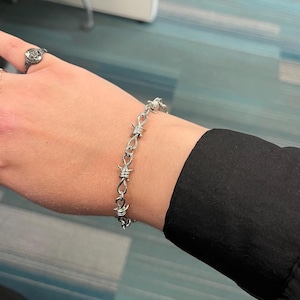 May include: Silver-toned bracelet featuring a barbed wire design. The bracelet is worn on a wrist, with a black sleeve visible in the background. A ring with a decorative design is on the index finger.
