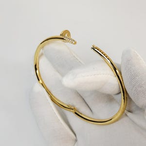 Puede incluir: Una pulsera dorada con diseño de cabeza de clavo. La pulsera presenta un diseño curvo y abierto con una pequeña sección de la cabeza del clavo cubierta de pequeñas piedras transparentes. La pulsera se muestra sobre un fondo blanco.