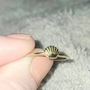 May include: A delicate gold ring featuring a seashell design. The ring has a thin band and a small, detailed seashell charm. The seashell is a light gold color with darker lines, creating a textured appearance. The ring is held between fingers.