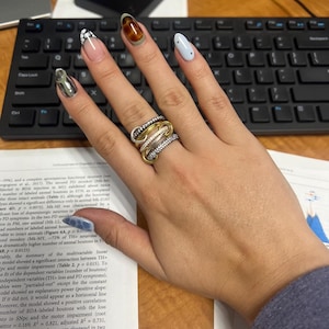 Op de afbeelding: Een hand met meerdere ringen met een goud, zilver en diamant ontwerp. De nagels zijn beschilderd met verschillende ontwerpen, waaronder zilver, bruin en lichtblauw. Een zwart toetsenbord is op de achtergrond.
