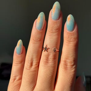 May include: A rose gold ring featuring a starfish and seashell design. The ring is worn on a finger with light blue nail polish with orange glitter accents. The ring is a delicate piece of jewelry.