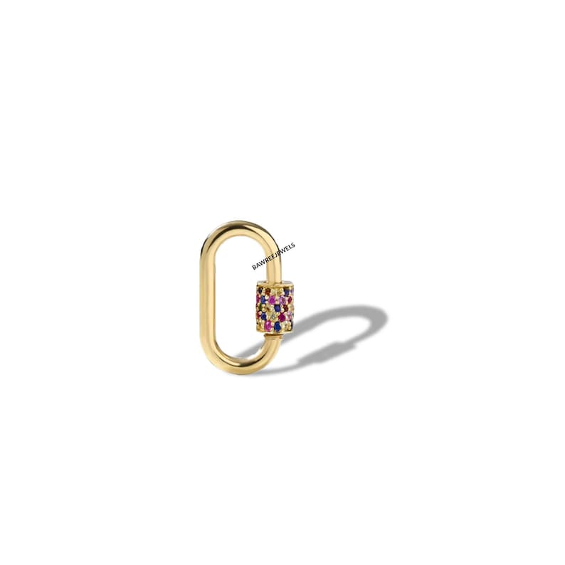 May include: A gold-coloured oval-shaped clasp with a cylinder of colourful gemstones. The clasp is a jewellery item, likely a pendant or charm. The gemstones are various colours, including pink, blue, and yellow. The clasp is on a white background.