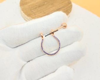 Puede incluir: Un anillo de septum en forma de herradura de color oro rosa con una hilera de pequeñas piedras azules a lo largo del borde curvo. La pieza metálica incluye una barra recta con un extremo esférico y el texto BAWREETEXTILES impreso en negro.