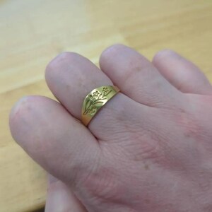 May include: A gold-colored ring with a floral design. The ring is worn on a finger and features an engraved pattern of leaves and flowers. The ring has a smooth, polished surface and a slightly curved shape.