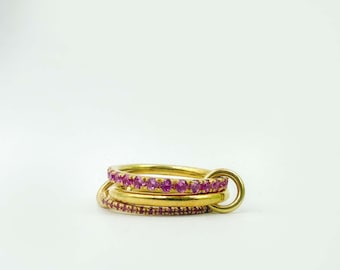 Può includere: Un set di tre anelli d'oro collegati tra loro. Gli anelli superiore e inferiore sono adornati con piccole e vibranti pietre preziose rosa. L'anello centrale è una fascia d'oro semplice. Gli anelli sono collegati da un anello d'oro.