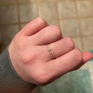 Puede incluir: Un delicado anillo de oro con un diseño en forma de cruz. El anillo es delgado y presenta pequeños acentos brillantes. El anillo se lleva en un dedo, mostrando sus intrincados detalles y su elegante simplicidad.