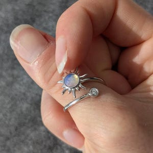 May include: A silver-colored ring featuring a sun design with a central iridescent stone and a small, clear gemstone. The sun has spiky rays, and the ring wraps around the finger with an open design.