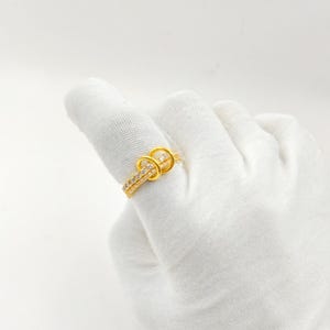 Op de afbeelding: Een goudkleurige ring met een knoopontwerp, bezet met kleine, heldere edelstenen. De ring bevindt zich op een vinger bedekt met een witte handschoen, tegen een witte achtergrond. Het ontwerp van de ring heeft twee in elkaar grijpende lussen.