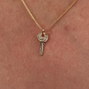 May include: A gold key pendant necklace. The key-shaped charm has a detailed design and hangs from a fine chain. The key has the text "APRIL IL CUORE" engraved on it. The necklace is displayed against a skin-toned background.
