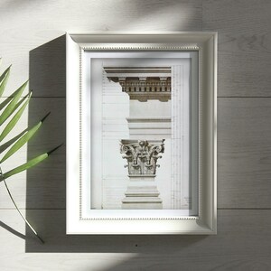 May include: A white framed print of a black and white architectural drawing of a column capital. The print is on a white wood surface with a green leaf in the foreground.