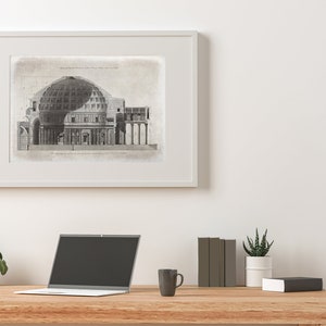 Puede incluir: Un dibujo arquitect&oacute;nico en blanco y negro de un edificio con una c&uacute;pula, enmarcado en un marco blanco y colgado en una pared blanca. Un port&aacute;til, una taza de caf&eacute; y dos plantas en maceta est&aacute;n sobre una mesa de madera delante del dibujo enmarcado.