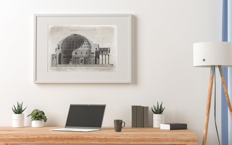 Puede incluir: Un dibujo arquitect&oacute;nico en blanco y negro de un edificio con una c&uacute;pula, enmarcado en un marco blanco y colgado en una pared blanca. Un port&aacute;til, una taza de caf&eacute; y dos plantas en maceta est&aacute;n sobre una mesa de madera delante del dibujo enmarcado.