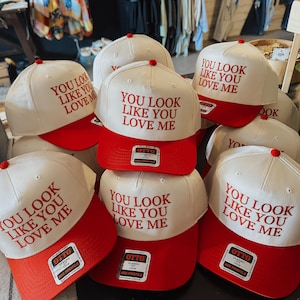 以下が含まれることがあります： 前面に赤い刺繍で「You look like you love me」と書かれた、白と赤のベースボールキャップが6つ。キャップは赤いつばと白いクラウンです。