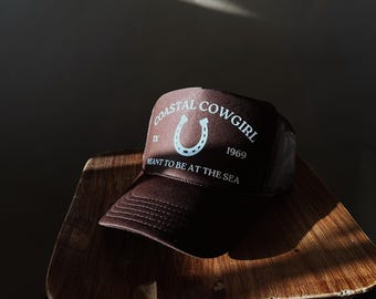Könnte beinhalten: Braune Trucker-Mütze mit dem Text "COASTAL COWGIRL" und einer Hufeisen-Grafik. Die Mütze trägt auch den Text "MEANT TO BE AT THE SEA" und "1969". Die Mütze liegt auf einem Holzschemel.