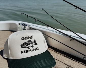 Puede incluir: Una gorra de béisbol blanca y verde oscuro con el texto "GONE FISHING" y un gráfico de pez. La gorra está en un barco con cañas de pescar en el fondo y una masa de agua.