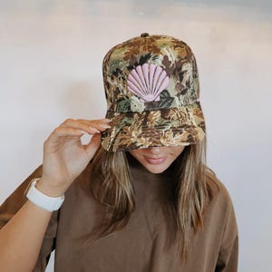 May include: A camouflage baseball cap with a pink seashell patch. The hat features a curved brim and a brown and green pattern. The seashell is a light pink color and is centered on the front of the cap.