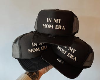 Könnte beinhalten: Schwarze Trucker-Caps mit weißem Text "IN MY MOM ERA". Die Caps haben einen Mesh-Rücken und einen gebogenen Schirm. Mehrere Caps sind übereinander gestapelt.