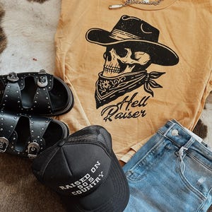 May include: An assortment of clothing and accessories, including a mustard yellow t-shirt with a skull wearing a cowboy hat and the text "Hell Raiser". Also, black sandals, a black trucker hat with "Raised on 90's Country" text, and blue jeans.