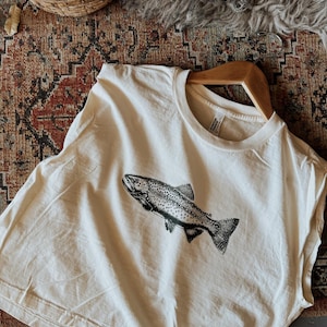 May include: Off-white t-shirt with a black and white fish illustration. The tee is displayed on a wooden hanger. A woven basket and patterned rug are visible in the background.