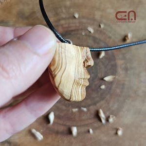 Puede incluir: Un colgante de madera tallado con la forma de un perfil humano, sujeto a un cordón negro. El colgante es de color marrón claro con vetas de madera visibles. El colgante está sujeto por una mano. El logotipo "CA Concetta Natale" está en la esquina superior derecha.