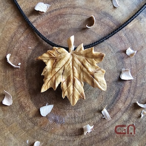 May include: A wooden maple leaf pendant necklace with a black cord. The leaf is carved with intricate details and has a natural wood finish.