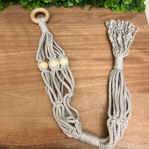 May include: A light grey macramé plant hanger with a wooden ring and three wooden beads. The plant hanger is made of twisted rope and has a knotted design. The bottom of the hanger has a tassel.