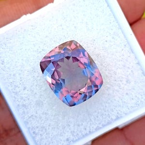 May include: A cushion-cut gemstone with a blend of pink, blue, and purple hues. The gem is set in a white, foam-like material, showcasing its facets and color variations. The stone's shape is rectangular with rounded corners.