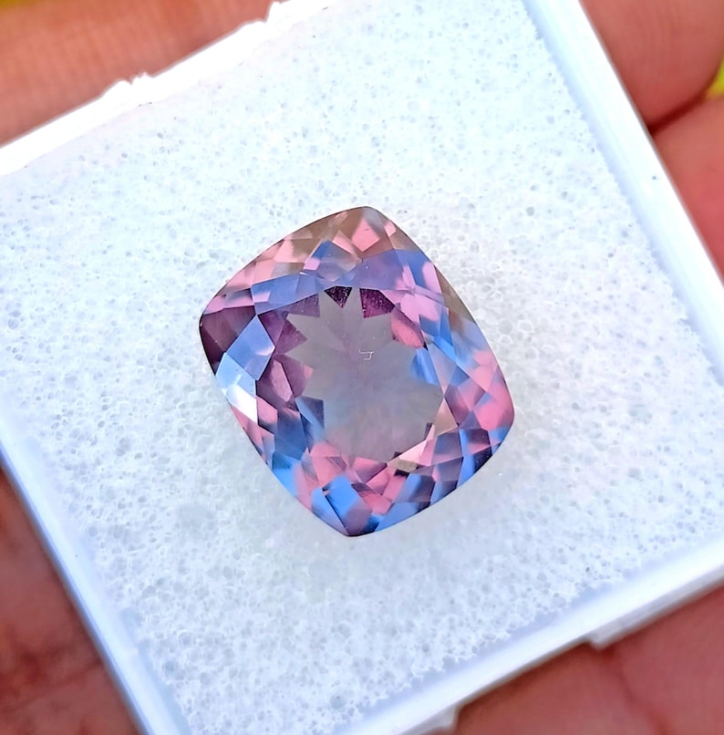 May include: A cushion-cut gemstone with a blend of pink, blue, and purple hues. The gem is set in a white, foam-like material, showcasing its facets and color variations. The stone's shape is rectangular with rounded corners.