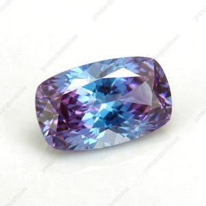 May include: A cushion-cut gemstone with a blend of blue and purple hues. The gem's facets reflect light, creating a shimmering effect. The gemstone is set against a white background.