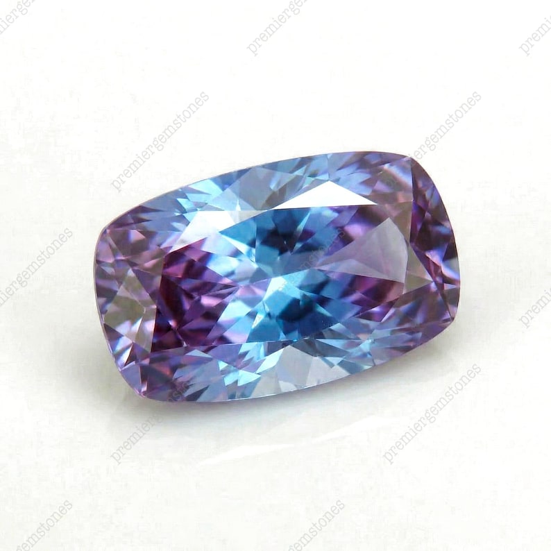 May include: A cushion-cut gemstone with a blend of blue and purple hues. The gem's facets reflect light, creating a shimmering effect. The gemstone is set against a white background.