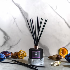 May include: A black diffuser bottle with black reeds and a copper-coloured collar. The bottle sits on a grey concrete base. The label reads "Plush Plum". The background includes plums and decorative elements.