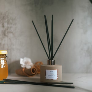 May include: A beige reed diffuser with black reeds and a gold-colored collar. The label reads "HONEY I'M HOME". A jar of honey and a rolled-up towel are in the background. The diffuser is a home fragrance product.