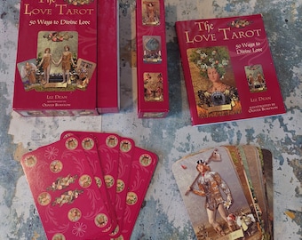 Puede incluir: Una caja roja con el título "The Love Tarot" y el texto "50 Ways to Divine Love". La caja contiene una baraja de cartas de tarot con ilustraciones de personas y escenas. Las cartas están dispuestas en forma de abanico. La caja está sobre una superficie texturizada azul y blanca.