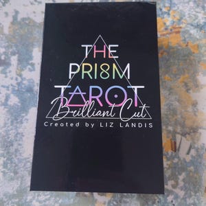 Op de afbeelding: De doos van het The Prism Tarot: Brilliant Cut deck heeft een zwarte achtergrond met een regenboogprisma-ontwerp en de tekst "The Prism Tarot: Brilliant Cut" en "Created by Liz Landis".