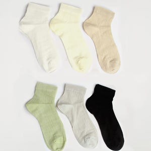 May include: Six pairs of ankle socks in various neutral colours are displayed on a white background. The socks include shades of white, cream, beige, light green, grey, and black. The socks have a textured, breathable knit.