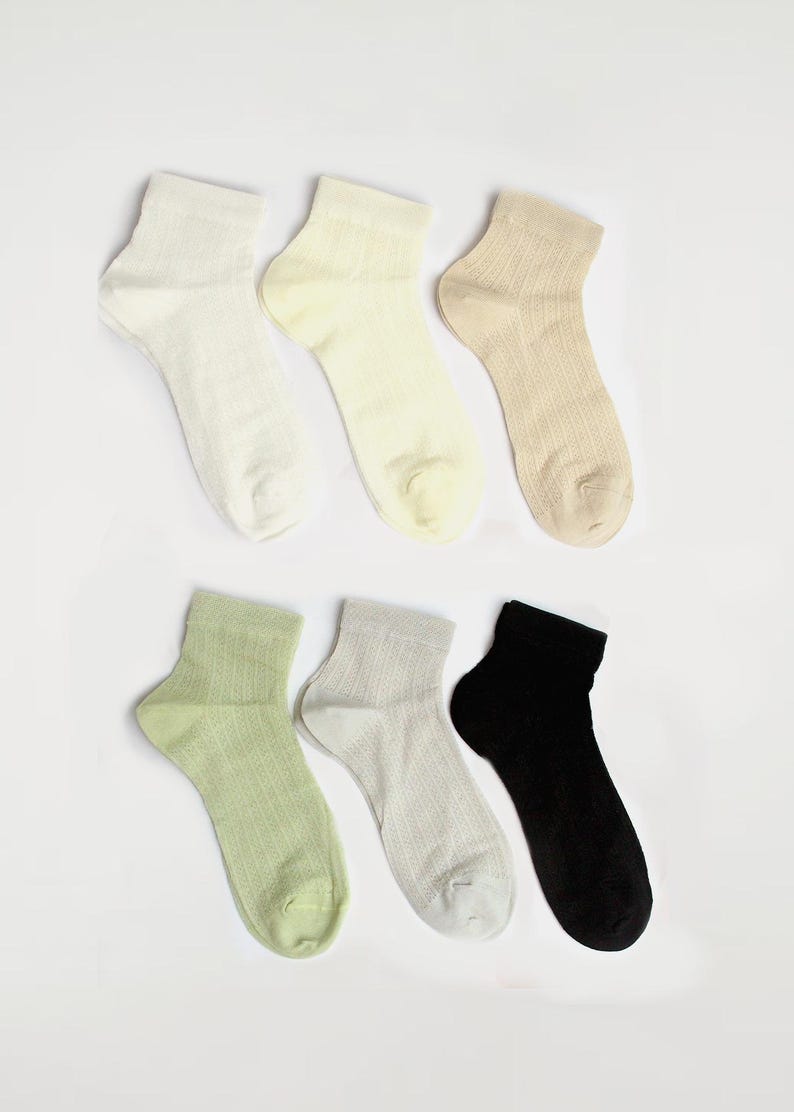 May include: Six pairs of ankle socks in various neutral colours are displayed on a white background. The socks include shades of white, cream, beige, light green, grey, and black. The socks have a textured, breathable knit.