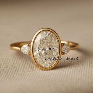 Puede incluir: Un anillo de oro con un diamante central de talla ovalada, engarzado en bisel, flanqueado por dos diamantes más pequeños en forma de pera. El anillo tiene una banda delicada y se muestra sobre una superficie de tono neutro. El texto "mehai gems" es visible.
