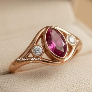 May include: A rose gold ring with a marquise-cut, deep pink gemstone as the centerpiece, flanked by two smaller, round, clear gemstones. The ring is set in a decorative, openwork design and rests in a beige ring box.