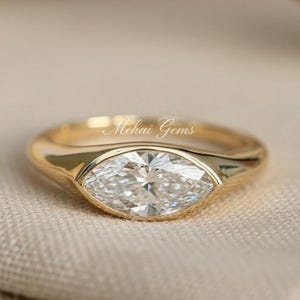 Könnte beinhalten: Ein goldener Ring mit einem Diamanten im Marquiseschliff. Der Ring hat ein glattes, poliertes Band und eine Lünettenfassung. Der Diamant ist klar und funkelnd. Der Ring liegt auf einem neutralfarbenen Stoff. Der Name Mehai Gems ist sichtbar.
