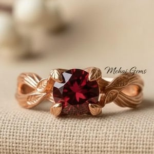 Puede incluir: Un anillo de oro rosa con una gema grande, redonda y de color rojo intenso. El anillo presenta un intrincado diseño de hojas en la banda y alrededor del engaste de la gema. El texto "Mehai Gems" es visible.