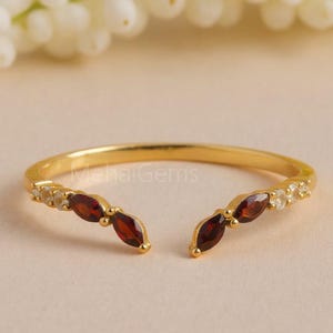 May include: A gold-colored ring with an open design, featuring alternating red and clear gemstones. The ring has a delicate band and is set against a soft, neutral background. The text "MehaiGems" is visible on the ring.