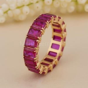 Könnte beinhalten: Ein goldfarbener Ring mit einem durchgehenden Band aus rechteckigen, tiefrosa Edelsteinen. Der Ring ist vor einem weichen, neutralen Hintergrund mit einem verschwommenen Blumenarrangement platziert. Der Text "Meha Gem" ist auf dem Ring sichtbar.