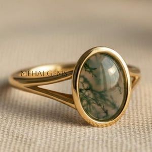 Könnte beinhalten: Ein goldener Ring mit einem ovalen Moosachat-Edelstein. Der Ring hat ein geteiltes Schaftdesign und der Text "MEHAI GEMS" ist auf dem Band sichtbar. Der Edelstein hat grüne und weiße Einschlüsse.